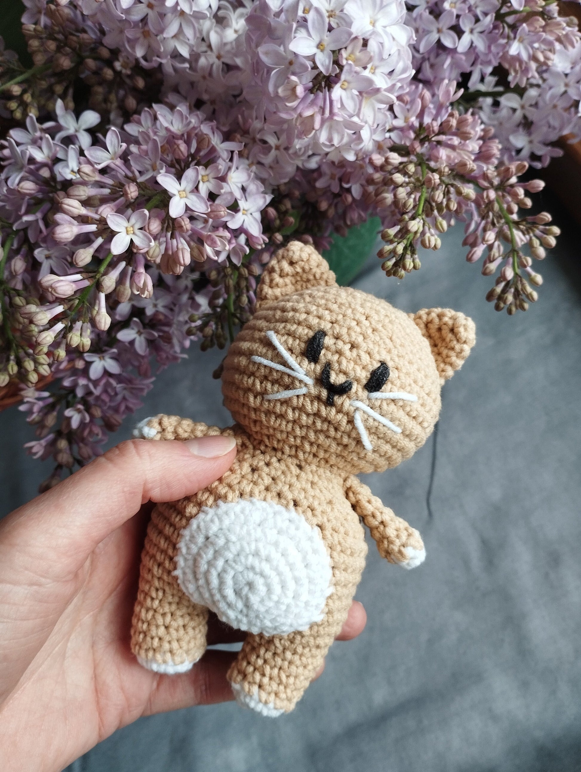 a hand holding a small crocheted cat next to a bouquet of flowers
