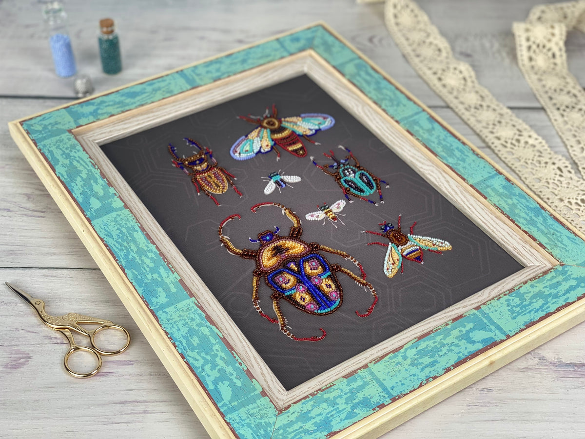 a framed artwork depicting various colorful insects, including a large dragonfly, on a dark background. The artwork is placed on a wooden surface, with a pair of scissors and some small bottles visible in the background.