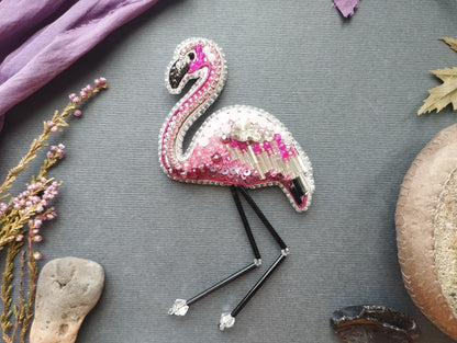 a pink flamingo embroidered on a grey background, surrounded by various decorative elements such as flowers, leaves, and a stone.