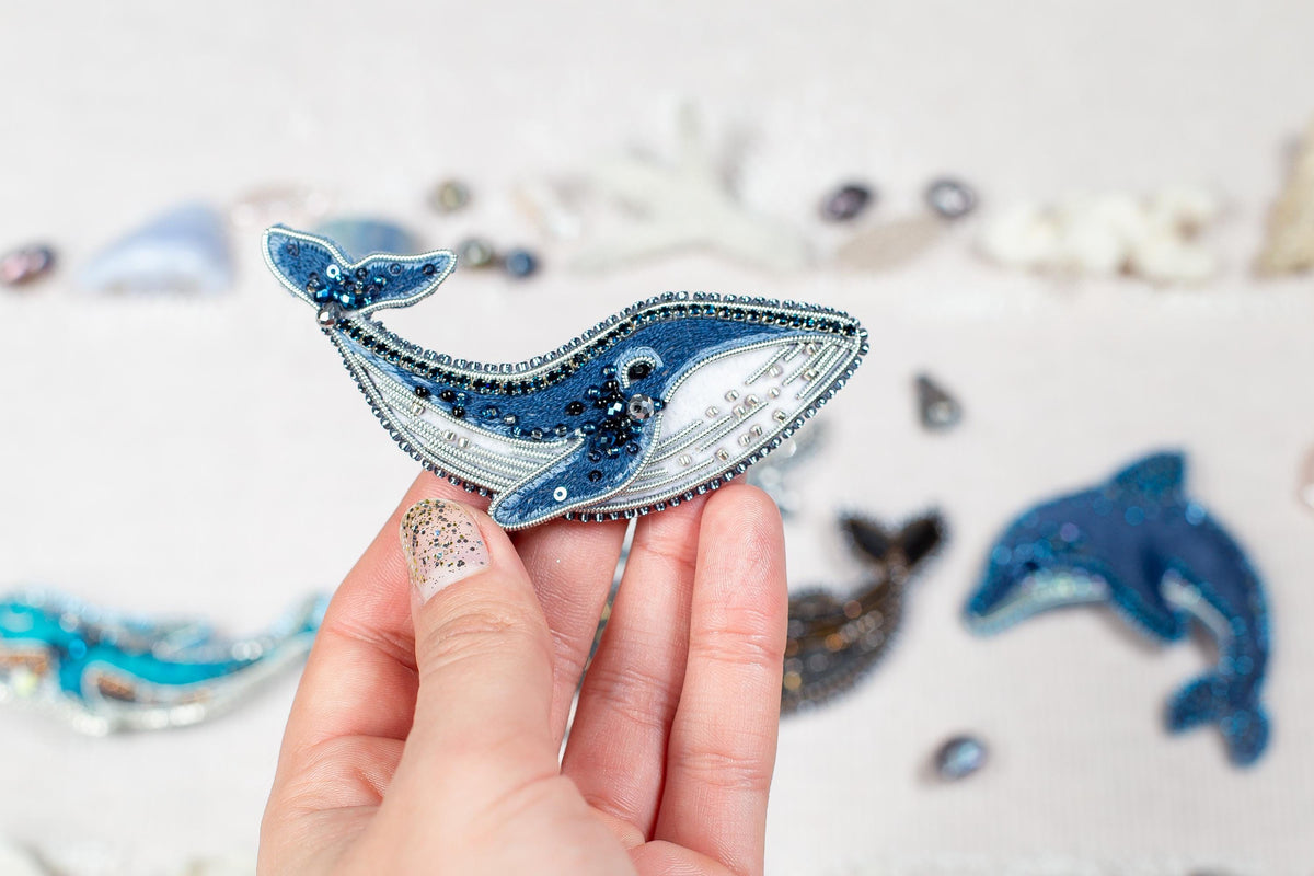 A hand holding a decorative blue and white beaded object resembling a whale or dolphin, with other similar objects in the background.
