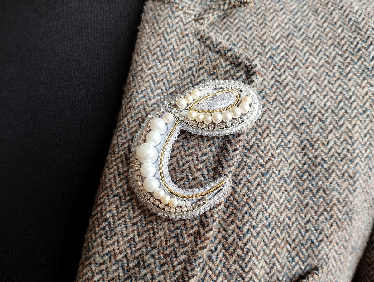 A close-up view of a decorative letter 'C' made of pearls and jewels, attached to a tweed fabric.