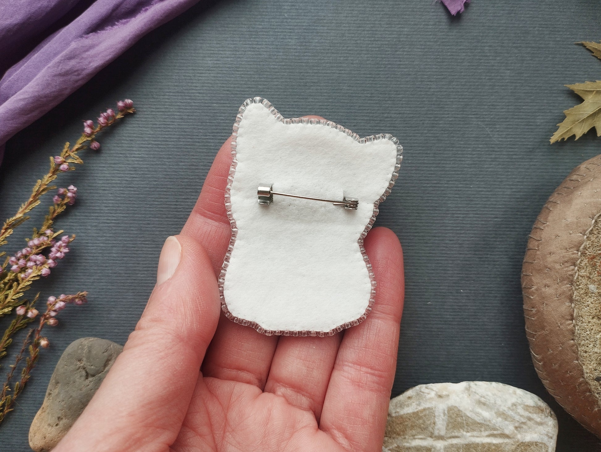 A hand holding a white felt cat pin against a dark background with various objects, including flowers and rocks.