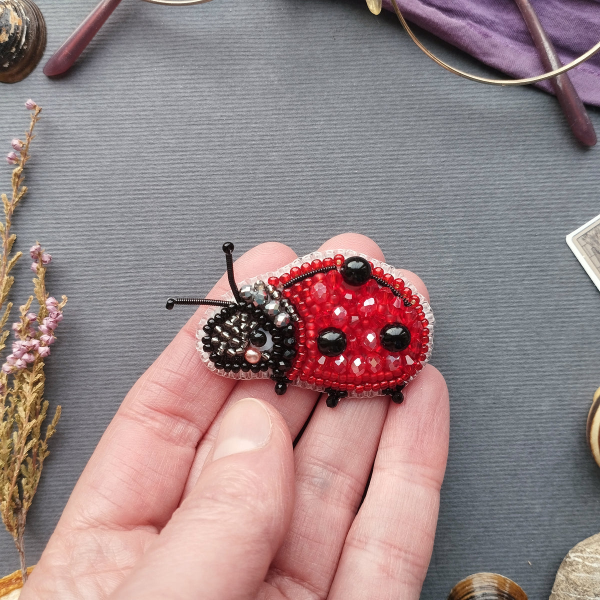 A hand is holding a small, intricately beaded ladybug brooch against a gray background.