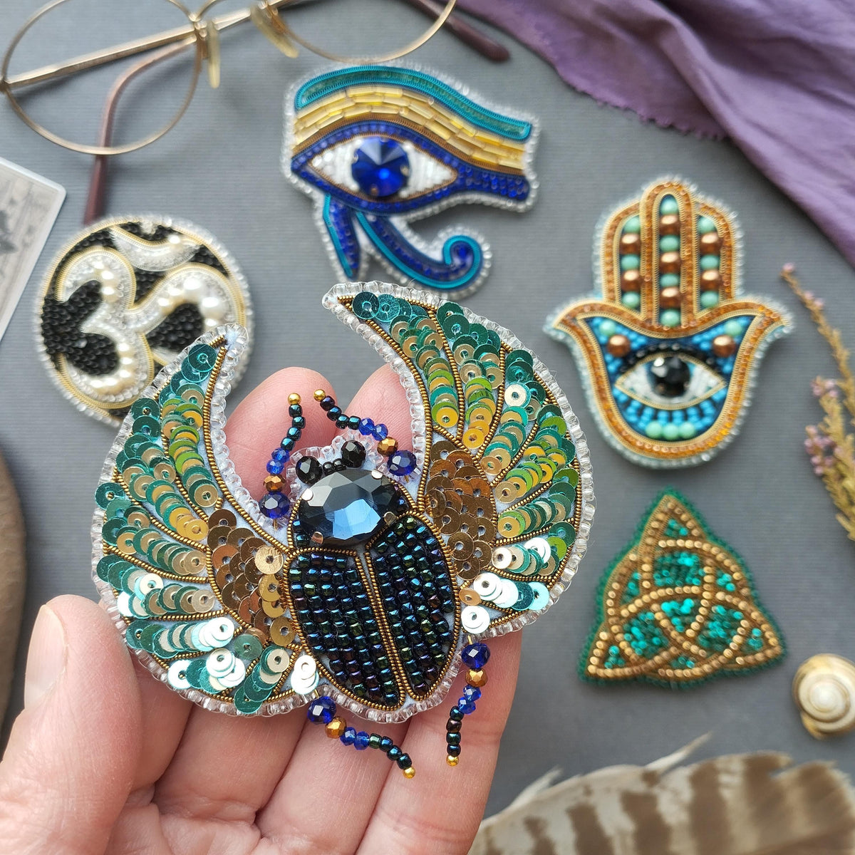 a hand holding a decorative piece of jewelry, which appears to be a beaded or embroidered design. The hand is holding a large, intricately designed piece that resembles a butterfly or a dragonfly. The background includes various other pieces of jewelry, such as a hamsa charm, a hand of faith charm, and a trident charm, all of which are also adorned with colorful beads and sequins.
