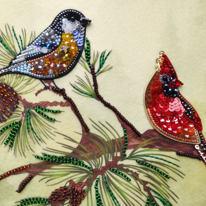 two birds, one black and one red, perched on a branch adorned with pine needles and pine cones. The birds are intricately crafted with beads and sequins, creating a detailed and vibrant scene.