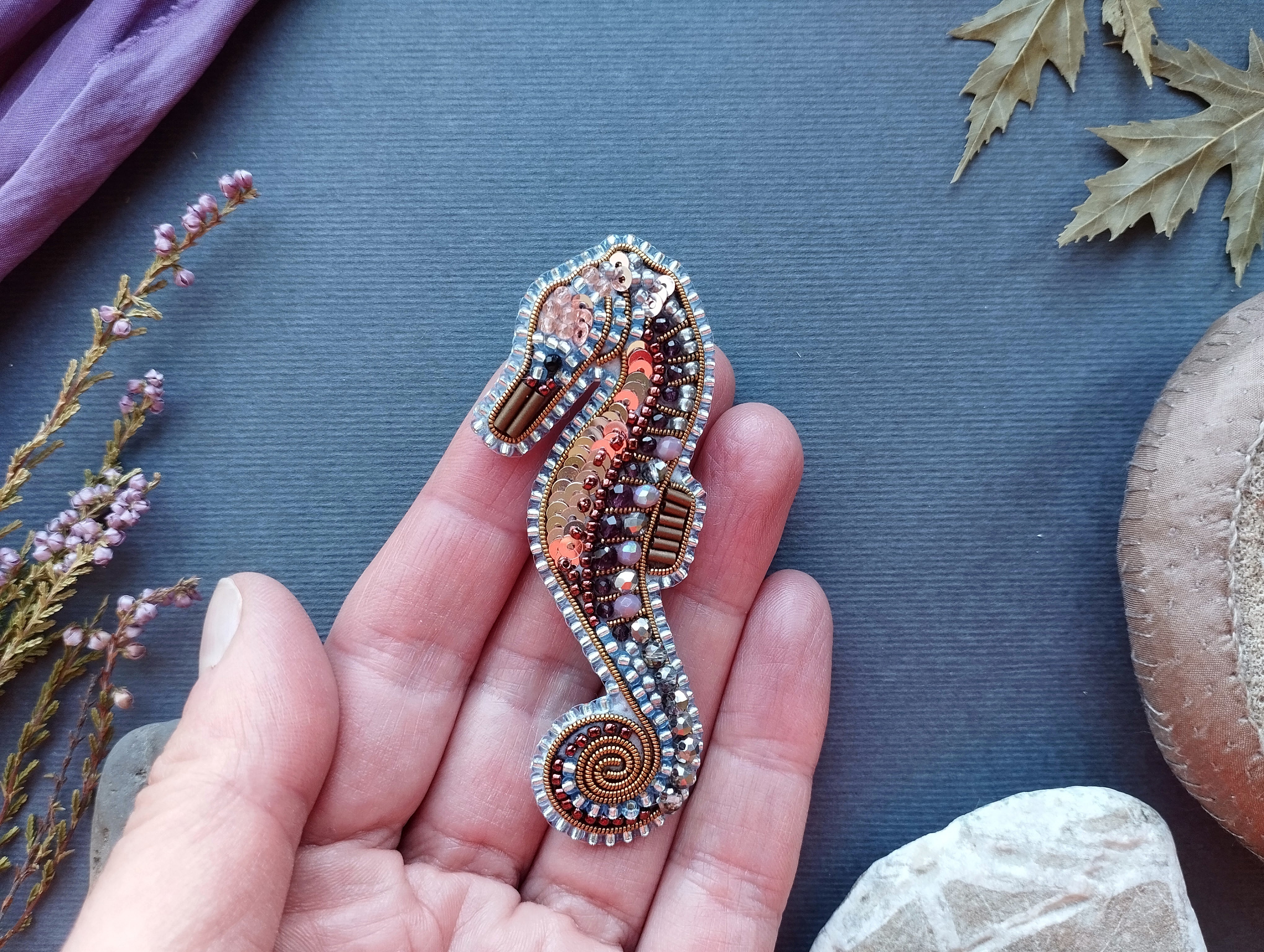 A hand holding a decorative beaded seahorse brooch against a blue background with some dried leaves and flowers.