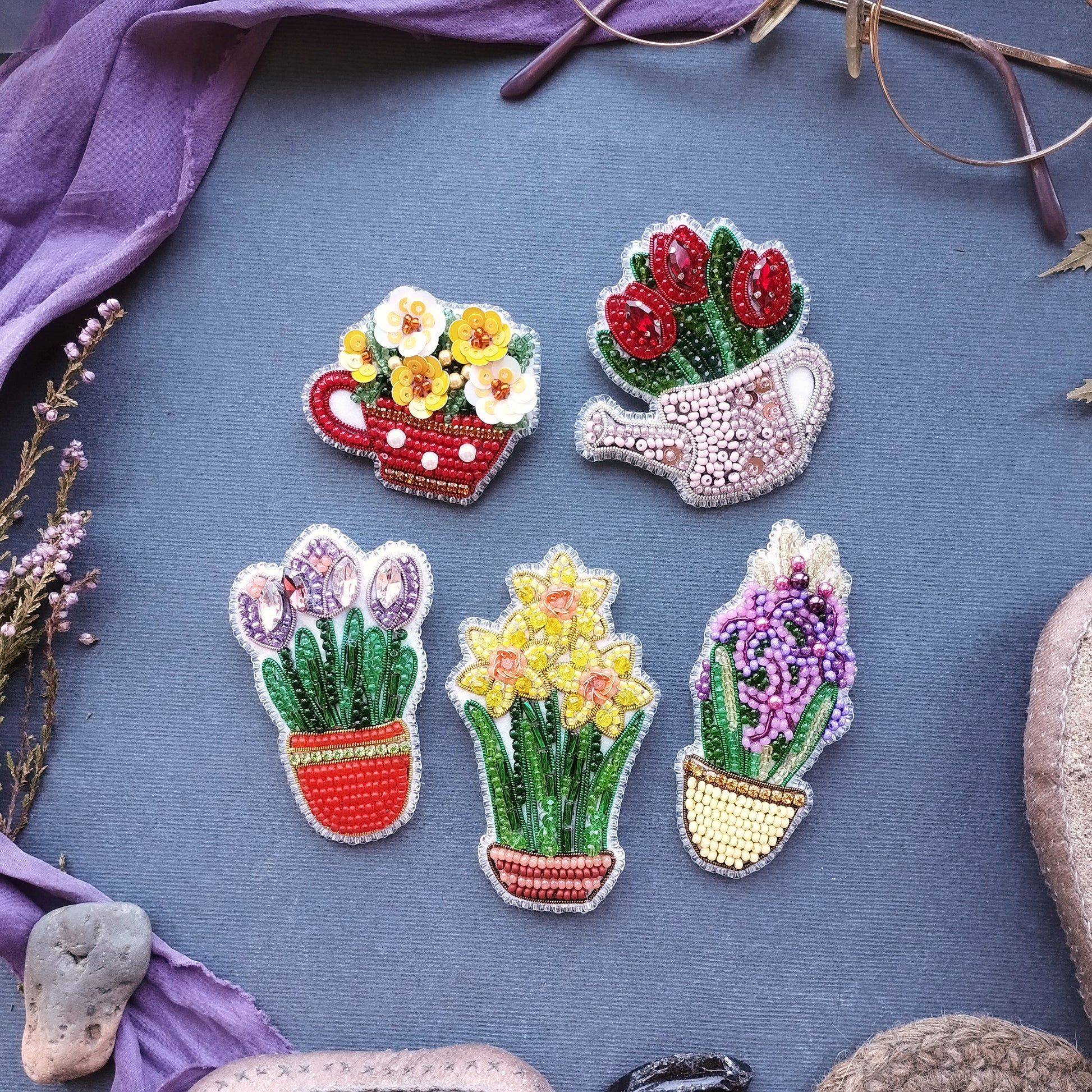 five decorative beaded brooches or pins, each depicting a different type of potted plant or flower, arranged on a blue background.