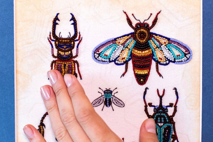 A hand is holding up a close-up view of a beaded artwork featuring colorful insects, including a large dragonfly and smaller flies.