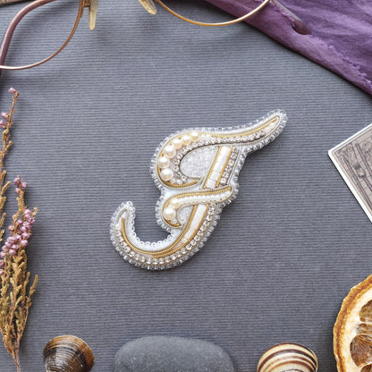 a decorative monogrammed letter "F" with intricate beading and pearl embellishments, surrounded by various objects such as a pair of glasses, a purple fabric, dried flowers, and a slice of orange.