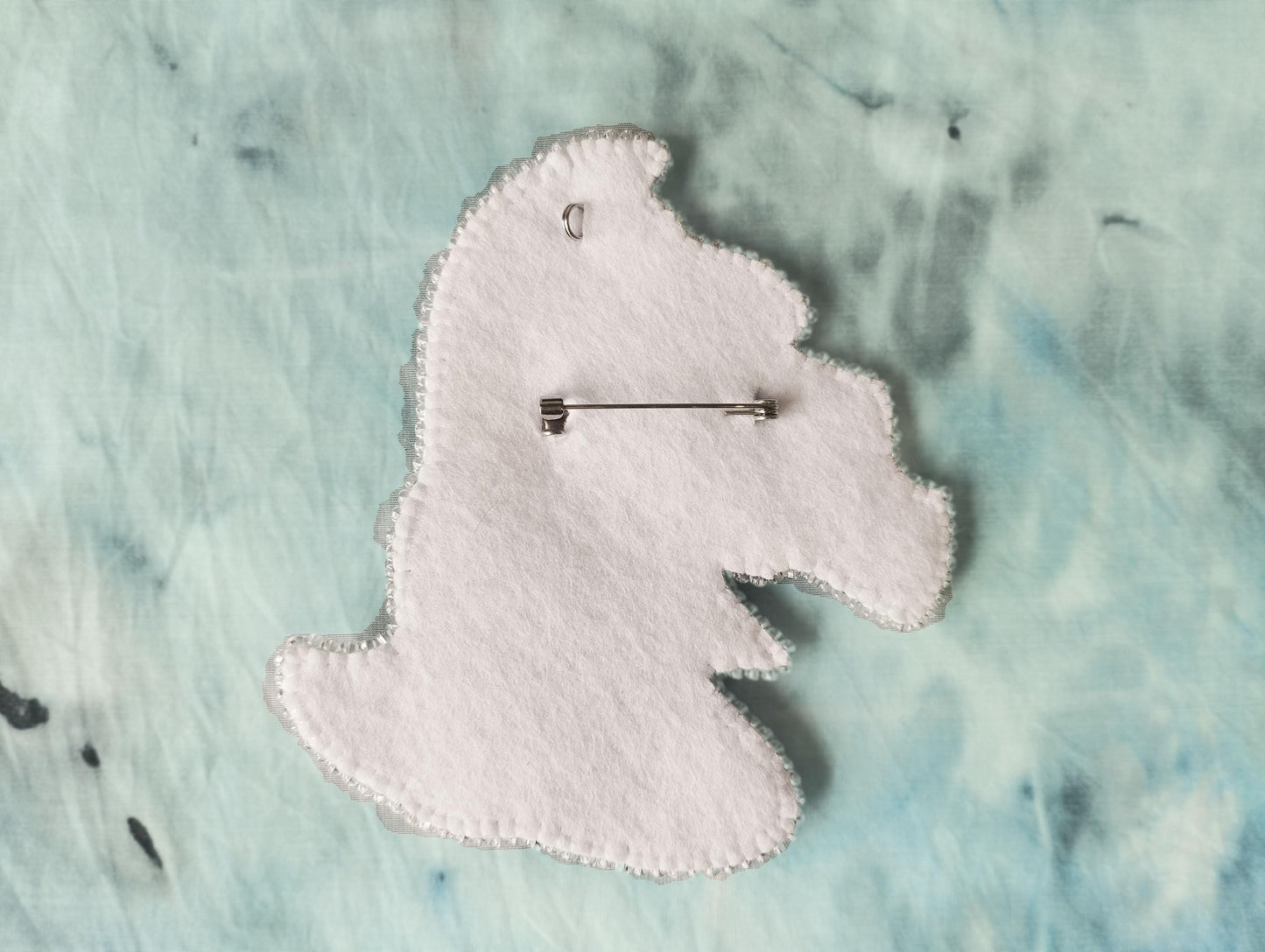 A white felt pin is attached to a fabric surface with a silver pinback.