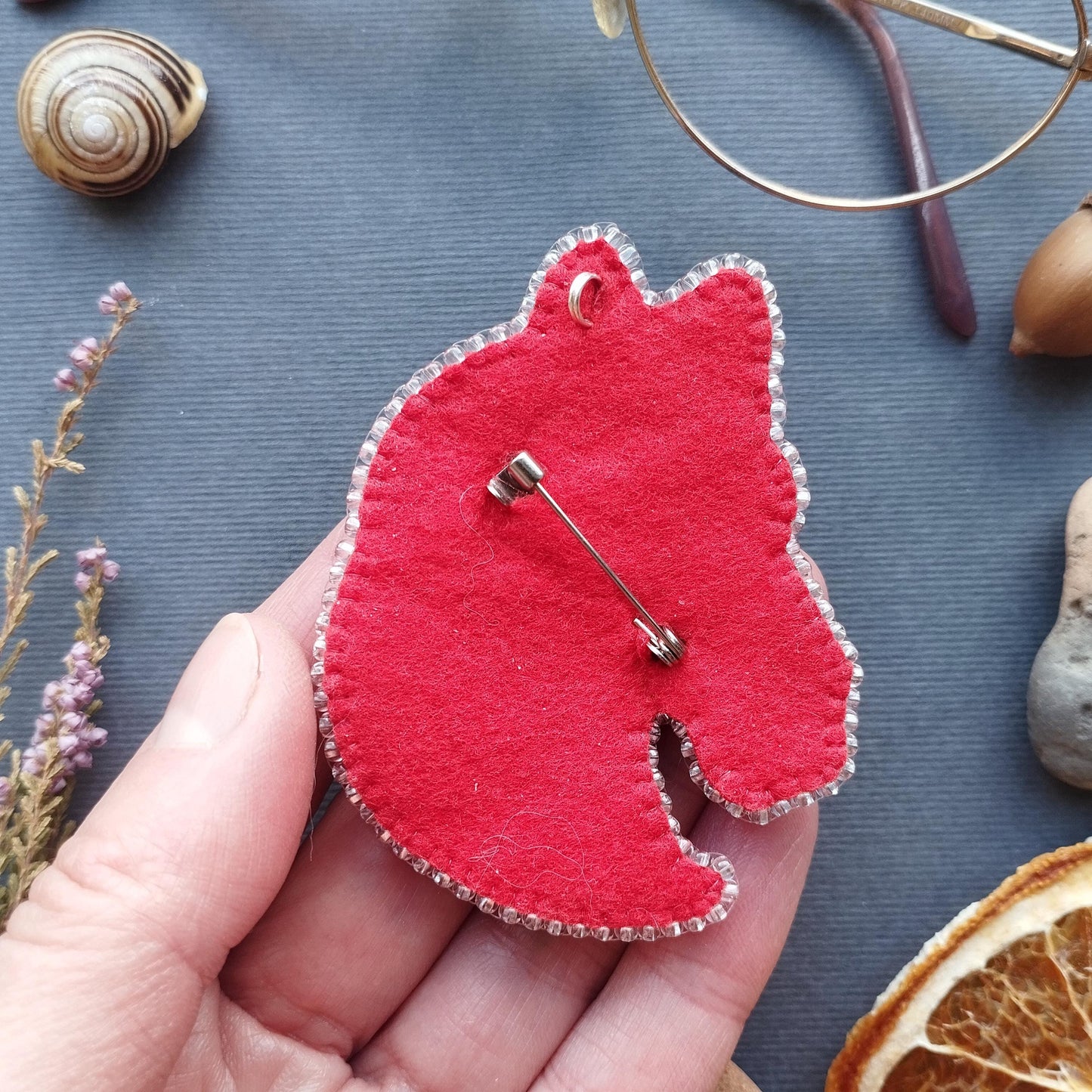 A hand holding a red felt keychain with a pin attached to it, surrounded by various objects such as a seashell, a pair of glasses, and dried flowers.
