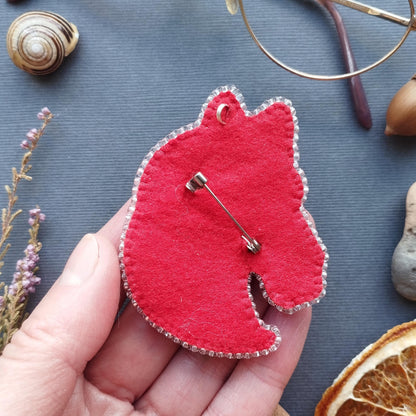 A hand holding a red felt keychain with a pin attached to it, surrounded by various objects such as a seashell, a pair of glasses, and dried flowers.