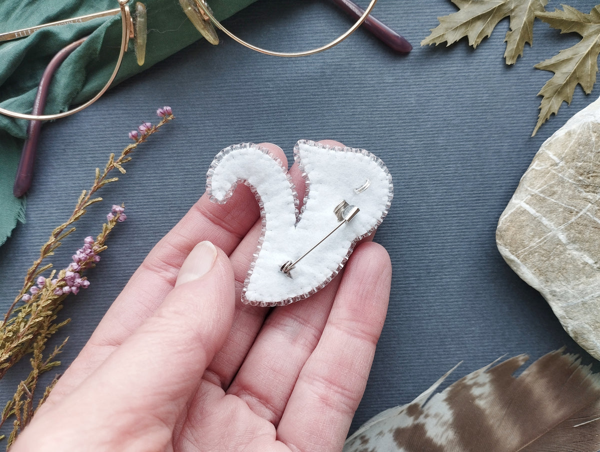 A hand holding a white felt object with a pin attached to it, surrounded by various natural elements such as leaves, feathers, and a pair of glasses.