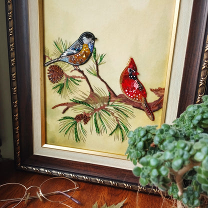a framed painting of two birds perched on a branch, surrounded by pine cones and pine needles. The painting is set against a wooden background and is framed in a gold frame. In the foreground, there is a potted plant and a pair of glasses on a wooden surface.