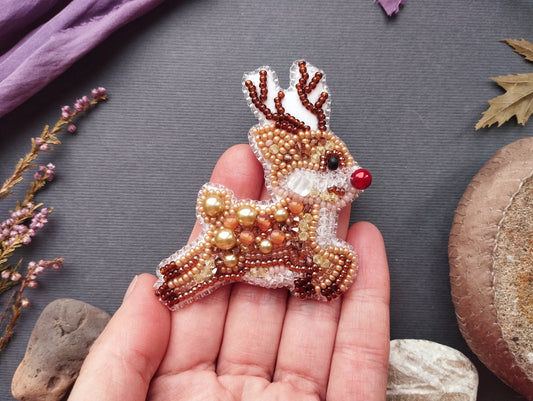 A hand holding a beaded reindeer brooch against a gray background.