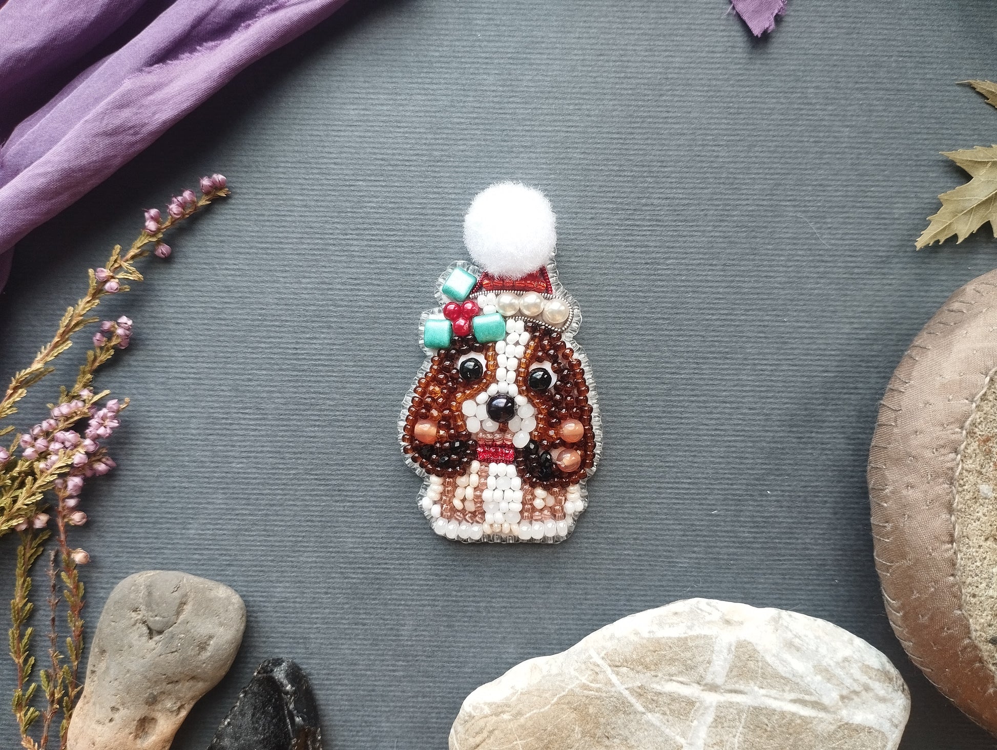 a small, intricately designed dog brooch made of beads and a pom pom, surrounded by various natural elements such as leaves, rocks, and flowers.