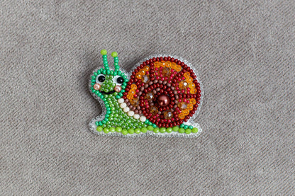 A colorful, hand-made beaded snail-shaped brooch or pin is displayed on a grey textured surface.