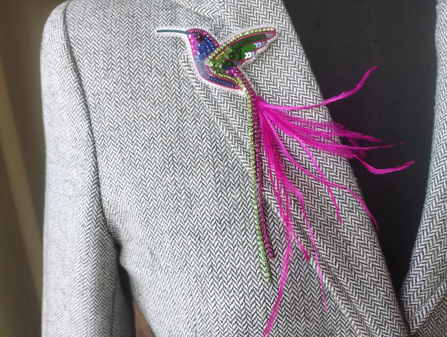 A close-up of a person wearing a gray tweed jacket with a colorful bird brooch on the left side of the jacket.
