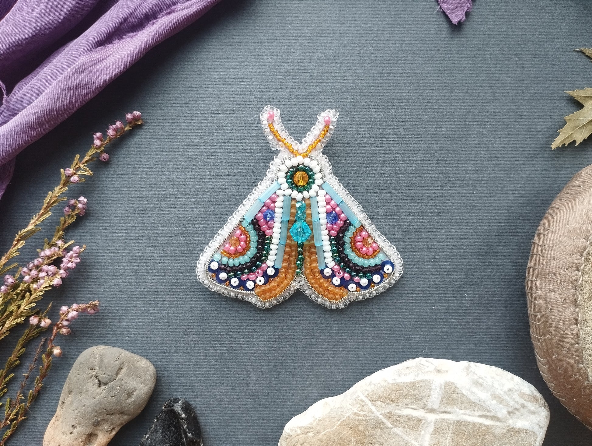 a colorful, hand-made butterfly brooch with intricate beading and beadwork, placed on a gray surface surrounded by various natural elements such as leaves, rocks, and flowers.