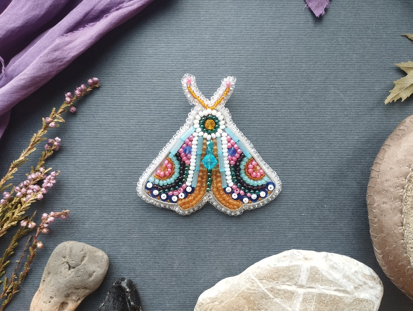 a colorful, hand-made butterfly brooch with intricate beading and beadwork, placed on a gray surface surrounded by various natural elements such as leaves, rocks, and flowers.
