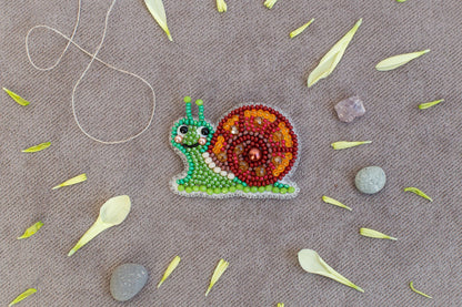 A colorful bead necklace depicting a snail is displayed on a beige surface, surrounded by scattered flower petals and small rocks.