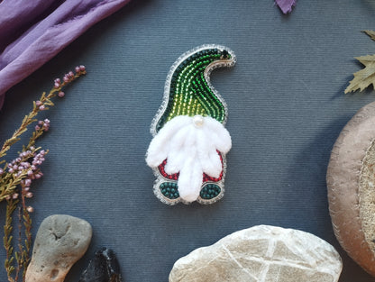 a small, intricately designed ornament resembling a gnome, with a white beard and green and red accents, placed on a dark surface surrounded by various natural elements such as leaves, rocks, and flowers.
