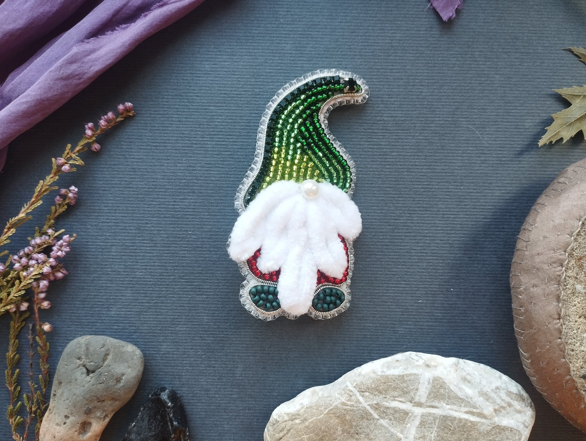 a small, intricately designed ornament resembling a gnome, with a white beard and green and red accents, placed on a dark surface surrounded by various natural elements such as leaves, rocks, and flowers.
