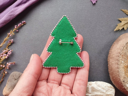 A hand holding a green felt Christmas tree pin against a gray background with various objects, including a purple fabric, leaves, and rocks.