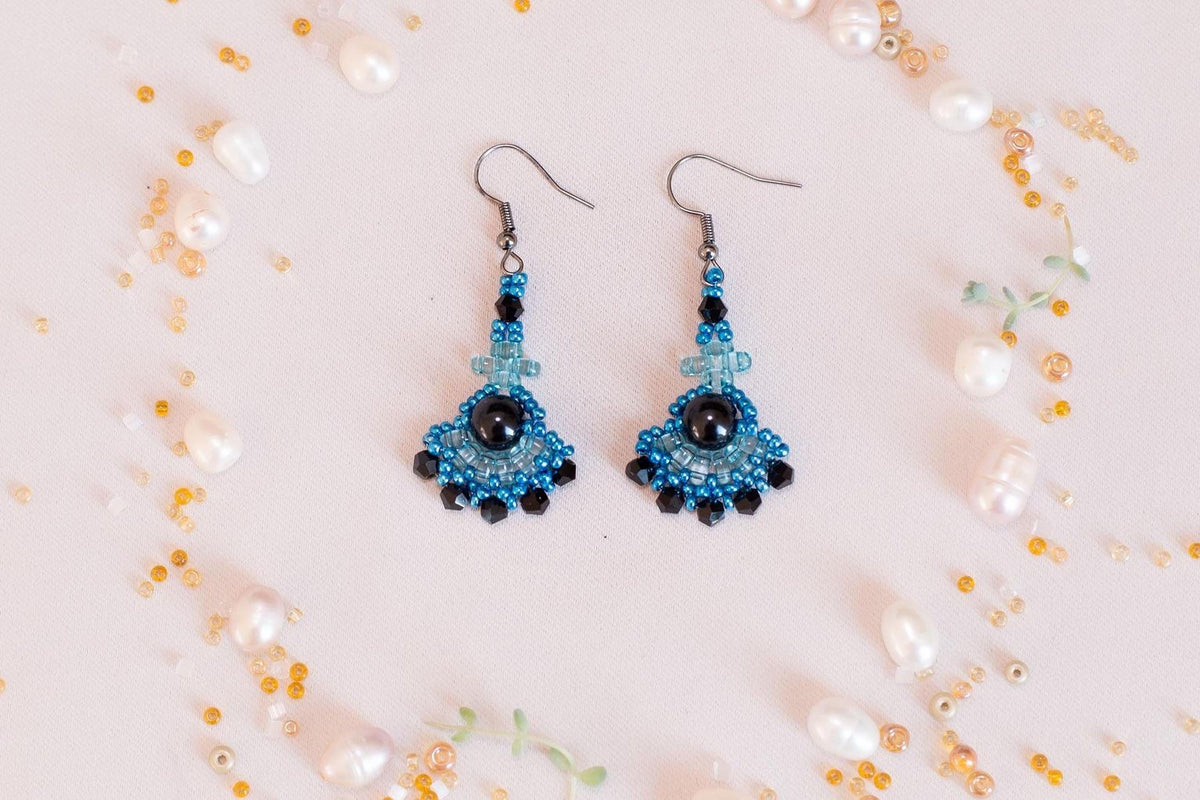 A pair of blue and black beaded earrings with a black gemstone is displayed on a light pink background, surrounded by scattered pearls and beads.