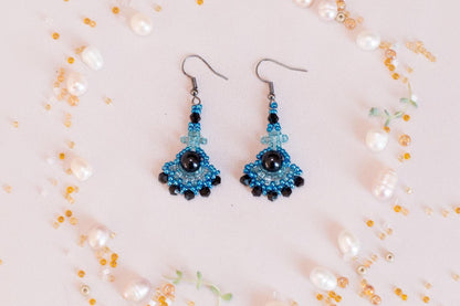 A pair of blue and black beaded earrings with a black gemstone is displayed on a light pink background, surrounded by scattered pearls and beads.