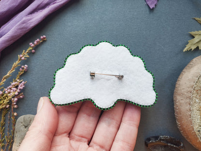 A hand holding a white felt pin with green accents, surrounded by various natural elements such as flowers, leaves, and a brown object.