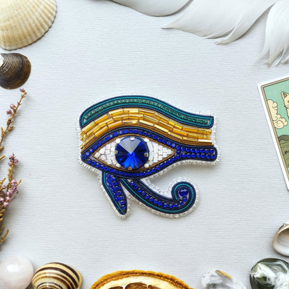a decorative eye-shaped piece of jewelry, adorned with blue and gold beads, and a large blue gemstone in the center. Surrounding the eye-shaped piece are various shells, feathers, and other natural elements, creating a visually appealing and harmonious composition.