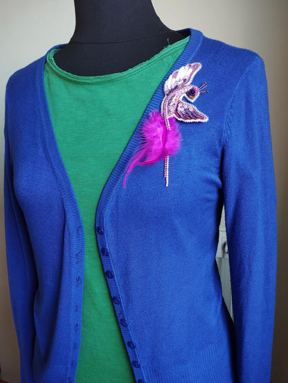 A mannequin wearing a blue cardigan with a green top underneath, and a pink feather brooch on the left side of the chest.