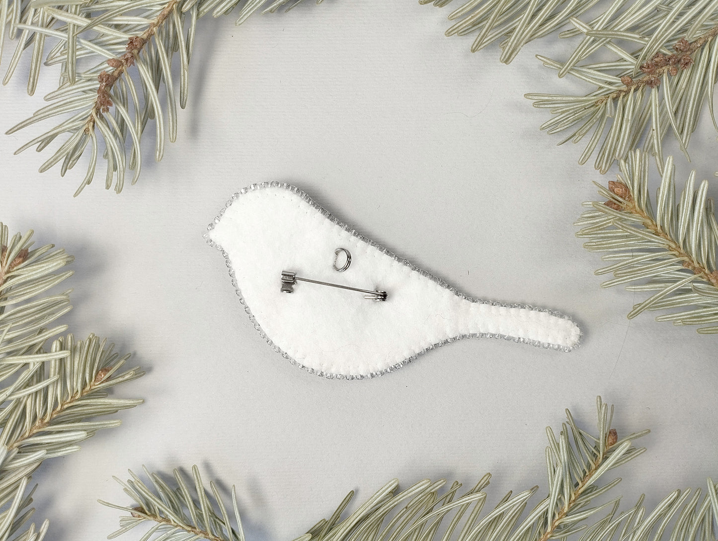 A white felt bird-shaped pin is surrounded by pine needles on a light background.