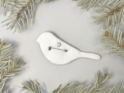 A white felt bird-shaped pin is surrounded by pine needles on a light background.