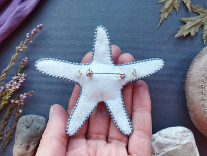 A hand holding a white starfish with blue accents, surrounded by various natural elements such as leaves, rocks, and flowers.
