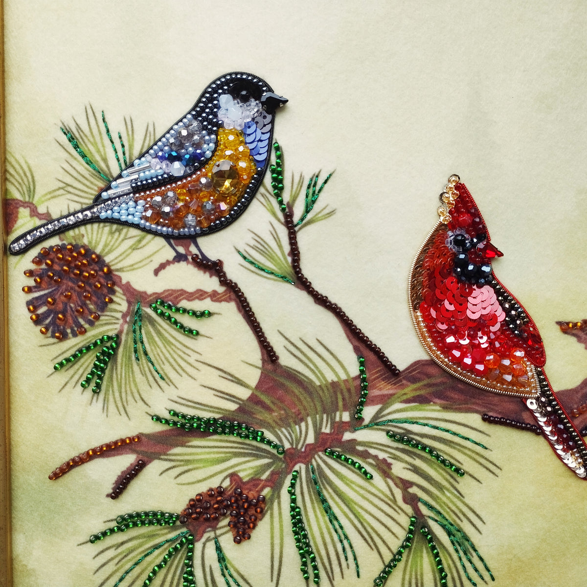 two birds, one red and one blue, perched on a branch with pine needles and pine cones. The birds are intricately detailed with beaded and embroidered elements, creating a vibrant and detailed representation of the natural world.