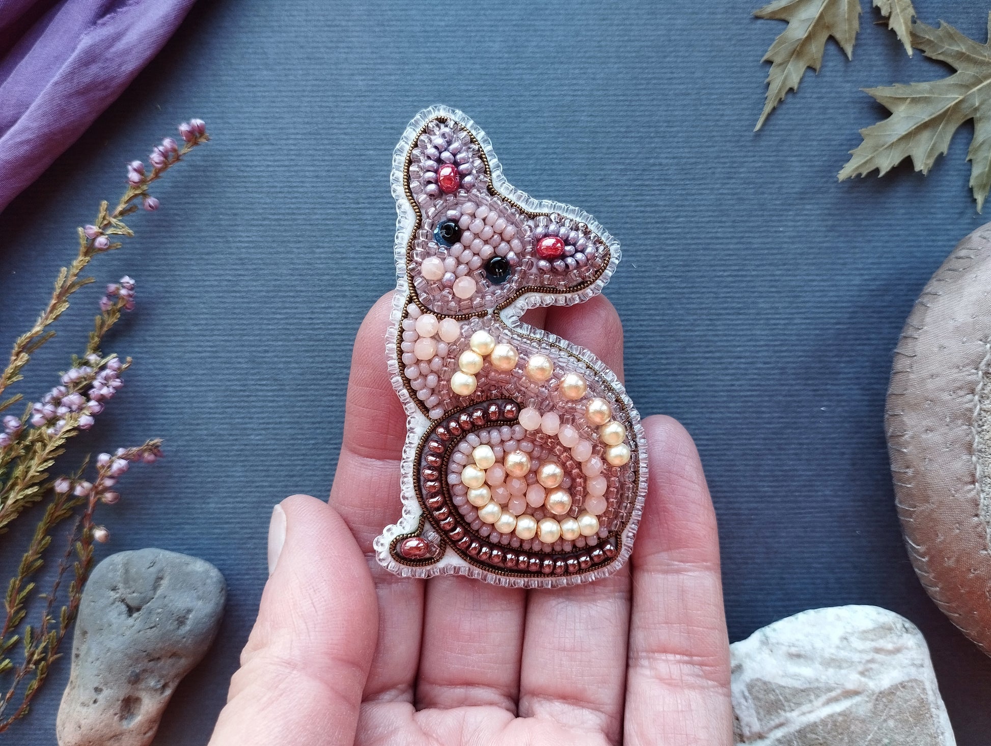 A hand holding a decorative beaded cat brooch against a blue background.