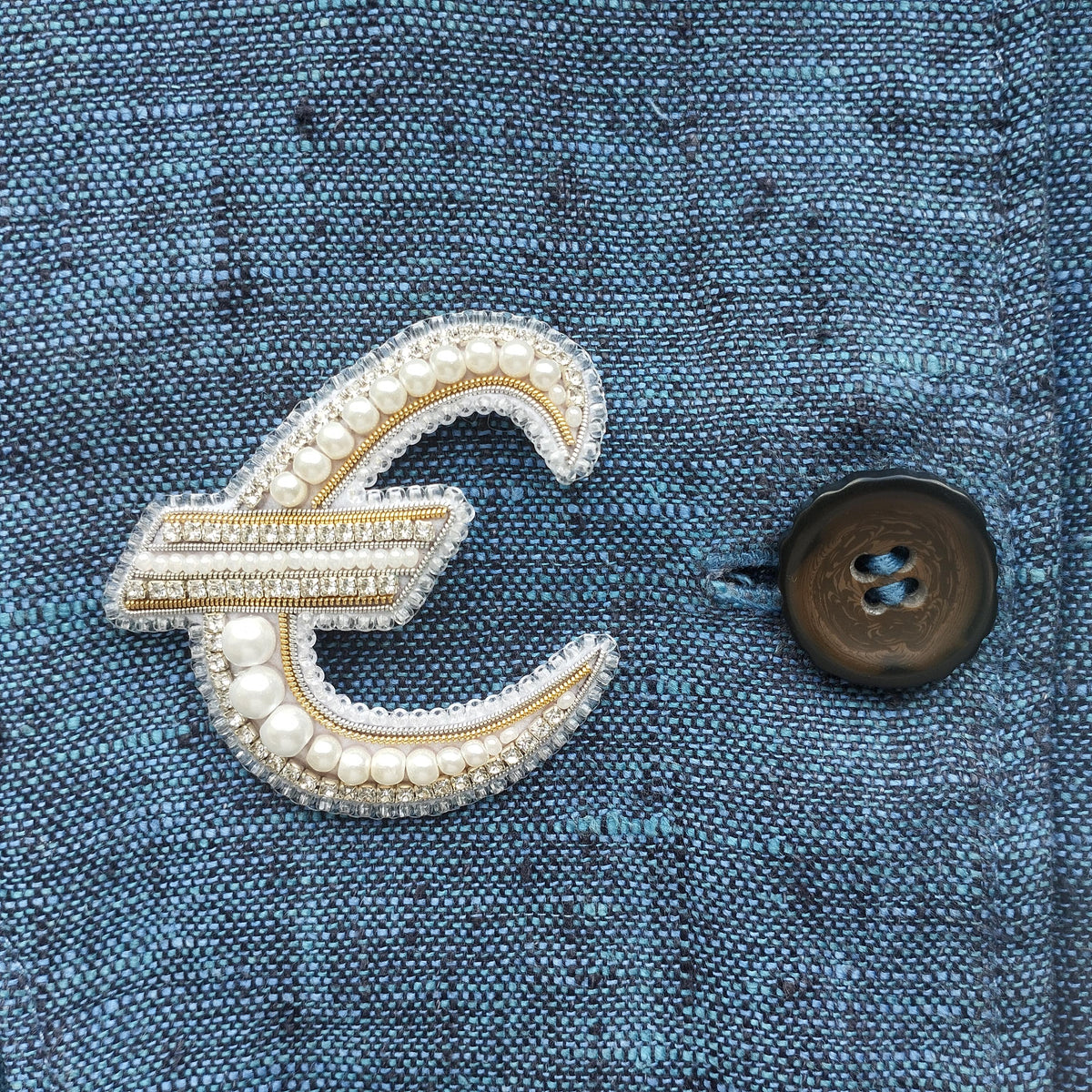 a close-up view of a blue fabric with a decorative letter 'E' made of pearls and rhinestones, and a button on the right side of the fabric.