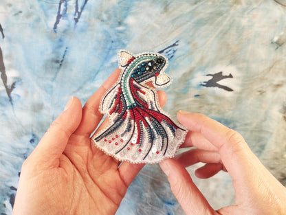 A hand holding a decorative beaded object resembling a fish or sea creature against a blue and white background.