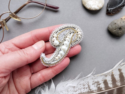 A hand holding a decorative pin with pearls and rhinestones, surrounded by various objects such as glasses, rocks, and feathers.