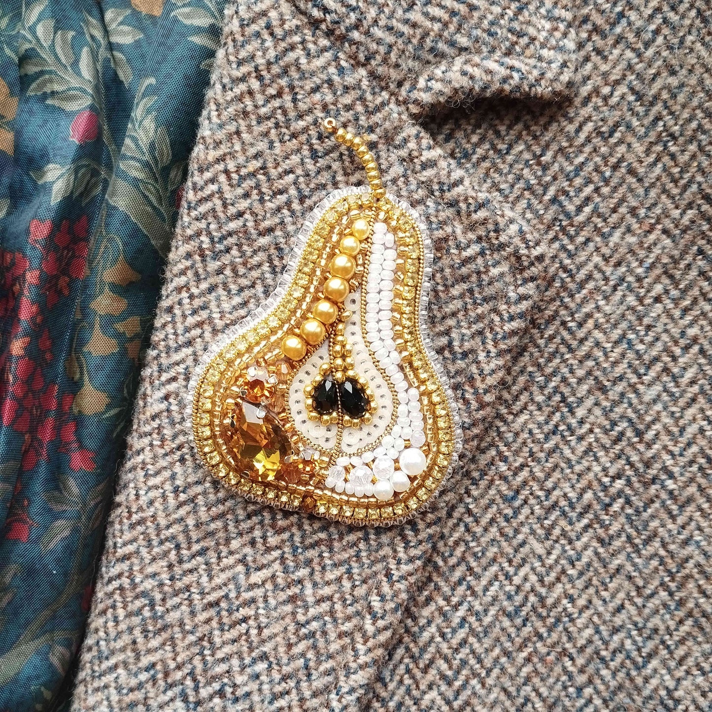 A close-up image of a decorative pin or brooch with a pear-shaped design, adorned with various embellishments such as beads, stones, and a gemstone.