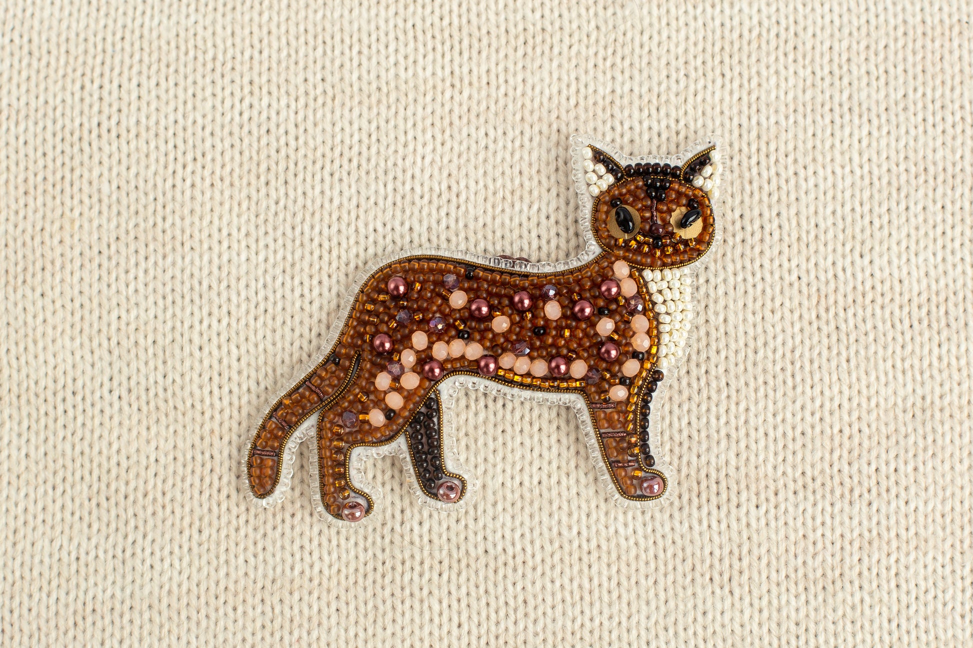 A decorative cat brooch made of beads and glitter, placed on a beige fabric background.