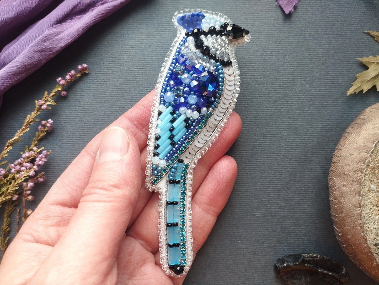 A hand is holding a hand-made beaded bird brooch against a dark background.