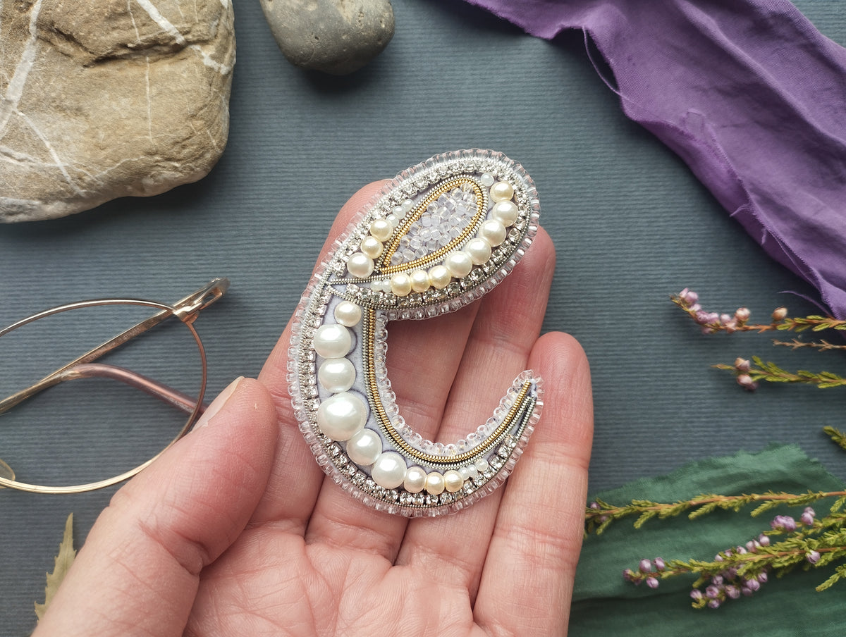 A hand holding a decorative letter 'C' made of beads, pearls, and crystals against a background of various objects, including rocks, flowers, and a purple fabric.