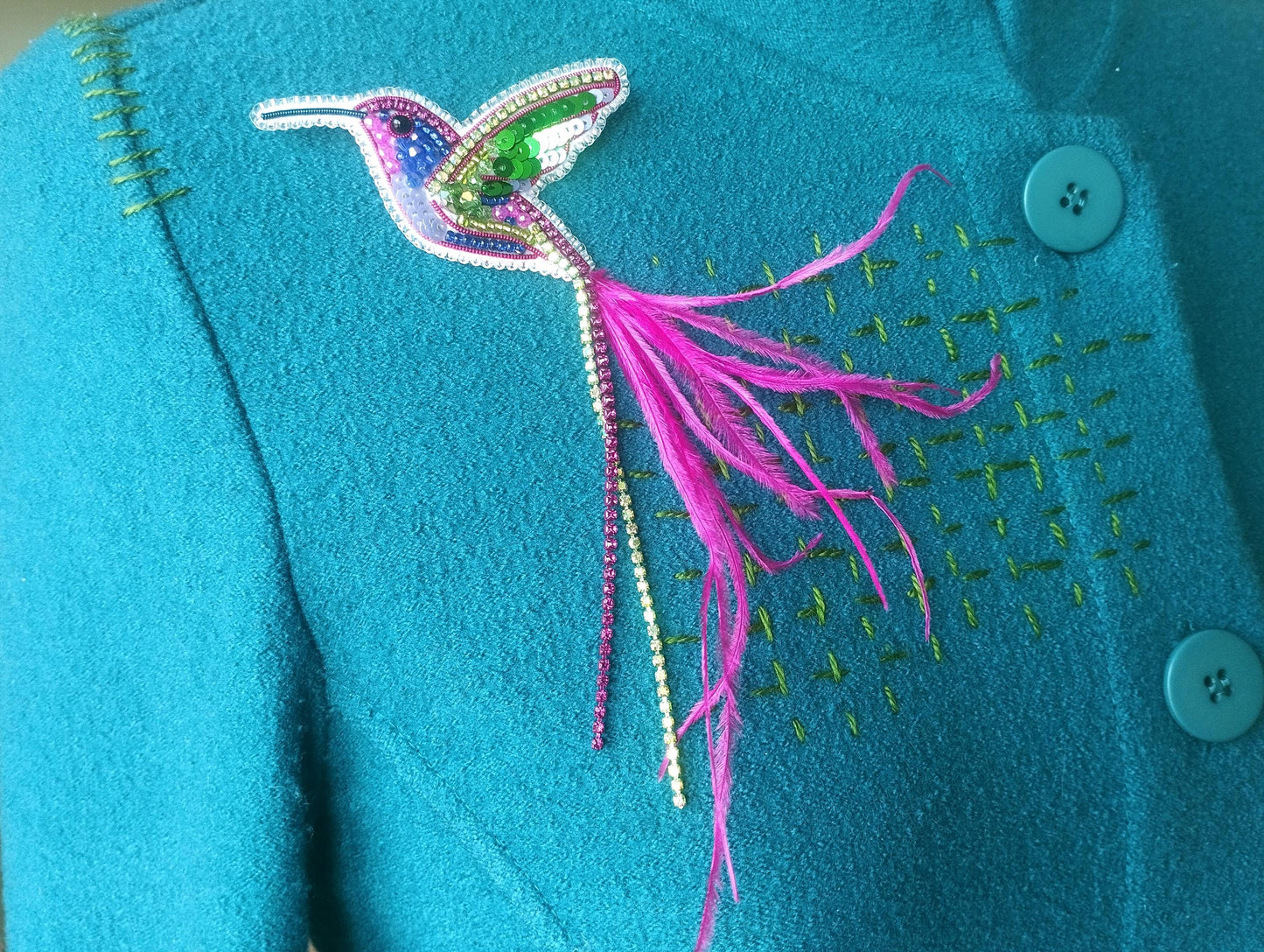 A close-up image of a teal-colored jacket with a decorative bird brooch featuring a colorful hummingbird design and a long pink feather.