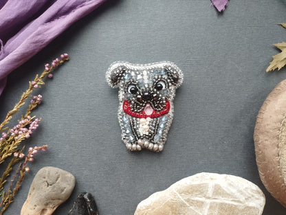 a decorative piece of jewelry, specifically a pug-shaped brooch, surrounded by various natural elements such as rocks, flowers, and leaves.