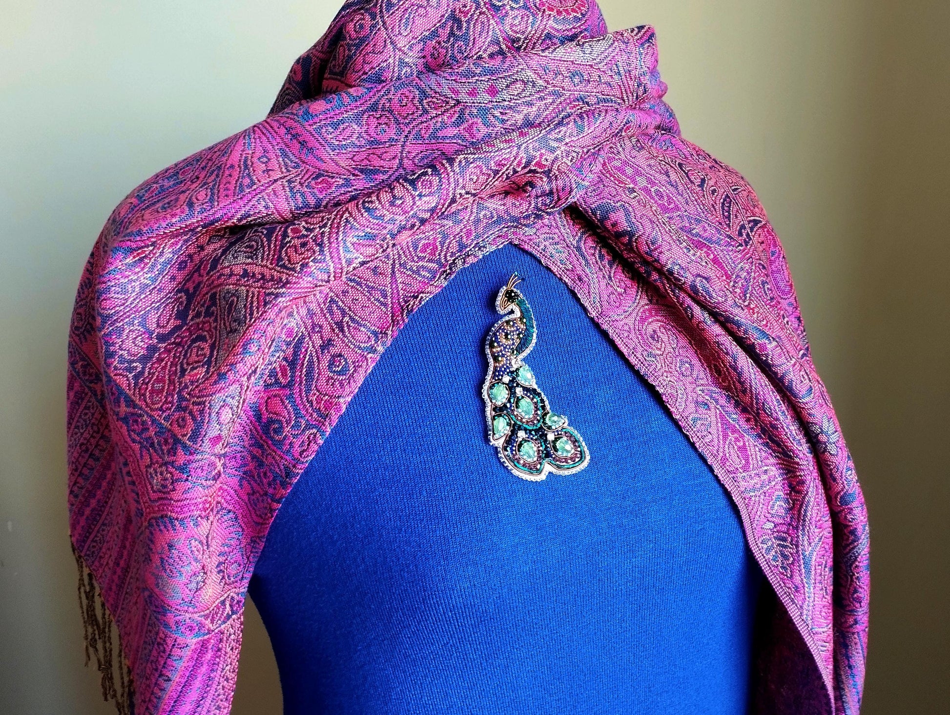A mannequin wearing a blue top with a pink and purple paisley patterned shawl draped over it, and a decorative brooch on the chest.