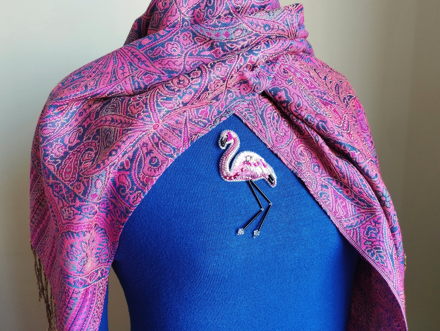 A mannequin wearing a blue top with a pink and purple paisley patterned scarf draped over it. The mannequin has a small, embroidered flamingo brooch pinned to the front of the blue top.