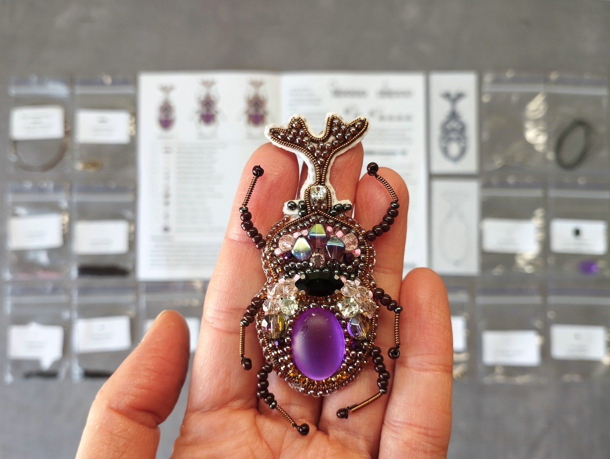A hand holding a decorative beaded insect-shaped brooch with a purple gemstone.